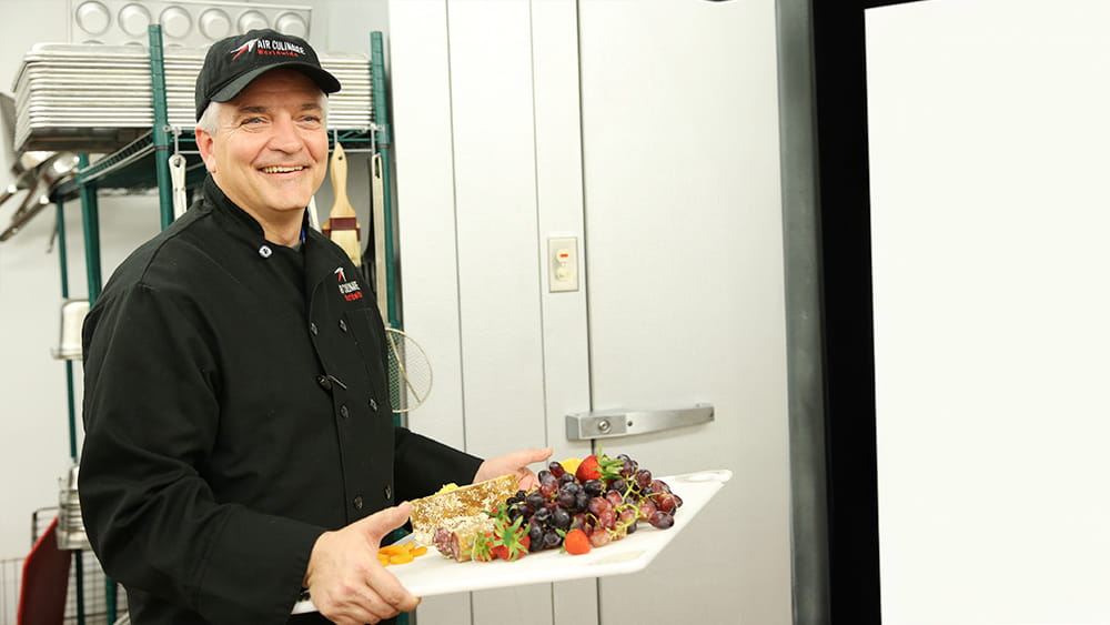 Air Culinaire Worldwide employee in chef uniform smiling holding a plate of fruit